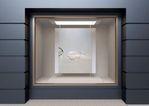 Muestra una vitrina en la pared con una escultura de cristal que tiene forma de dos medias esferas. La estructura de la vitrina es moderna y minimalista, con un marco en color gris oscuro.